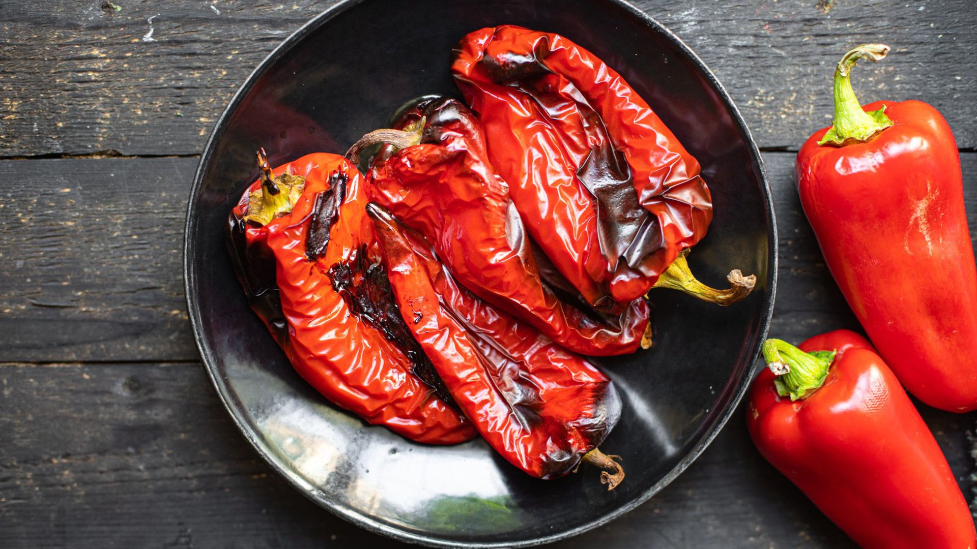 kako očistiti kožicu s pečenih paprika crna zdjela sa četiri pečene crvene paprike roge pored dvije sirove paprike crvene na podlozi od tamnih daski