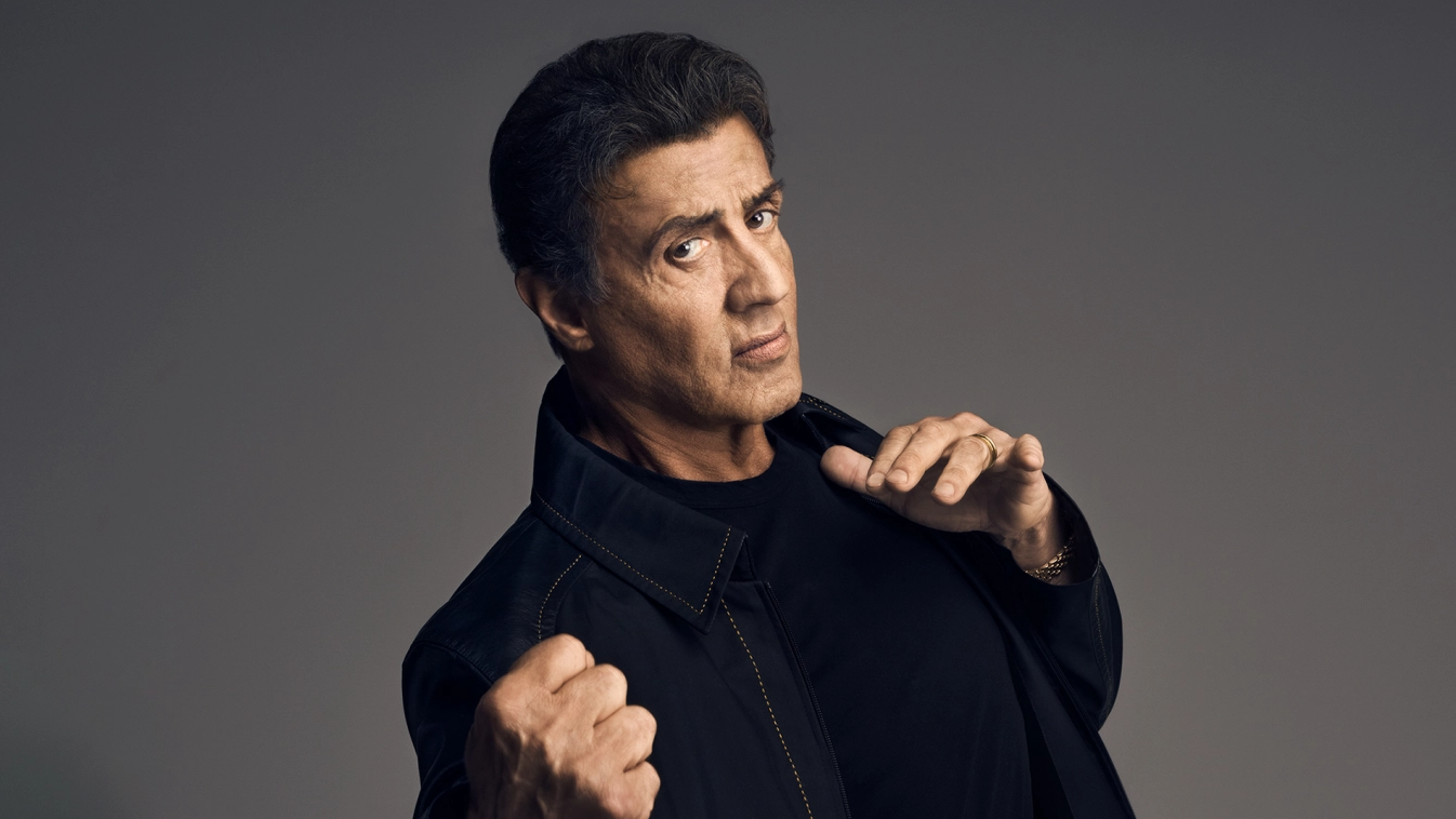 Sylvester Stallone u crnoj jakni i crnoj majici vidi se torzo desna šaka stisnuta lijeva ispružena na lijevoj ruci zlatni prsten na prstenjaku i zlatni sat siva podloga iza njega