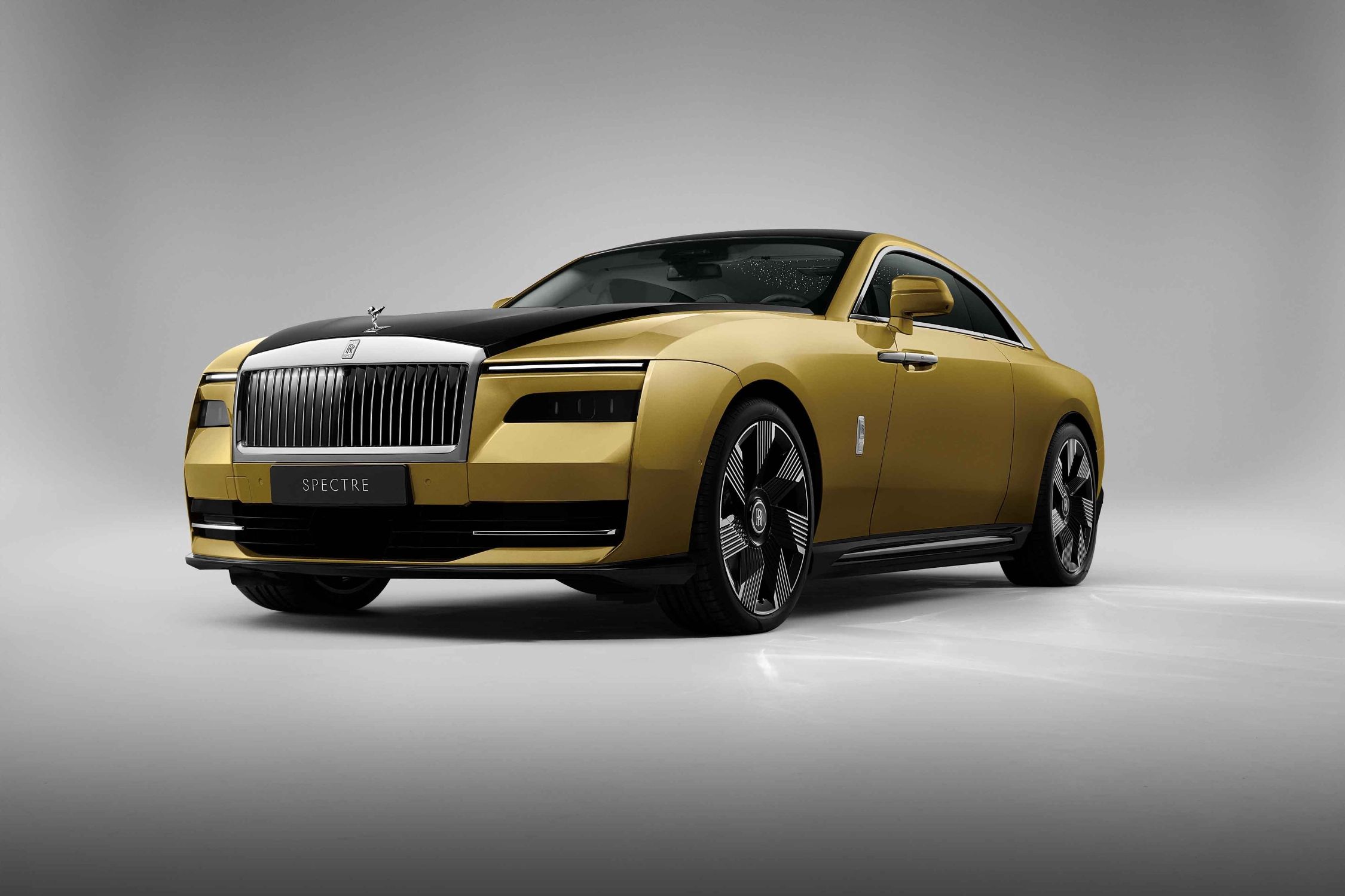 Rolls-Royce novi model Spectre automobil zlatne i crne boje crna boja na dijelu prednje haube i prati liniju preko krova koji je potpuno crn i gepeka siva podloga iza automobila kao daq je u prostoriji