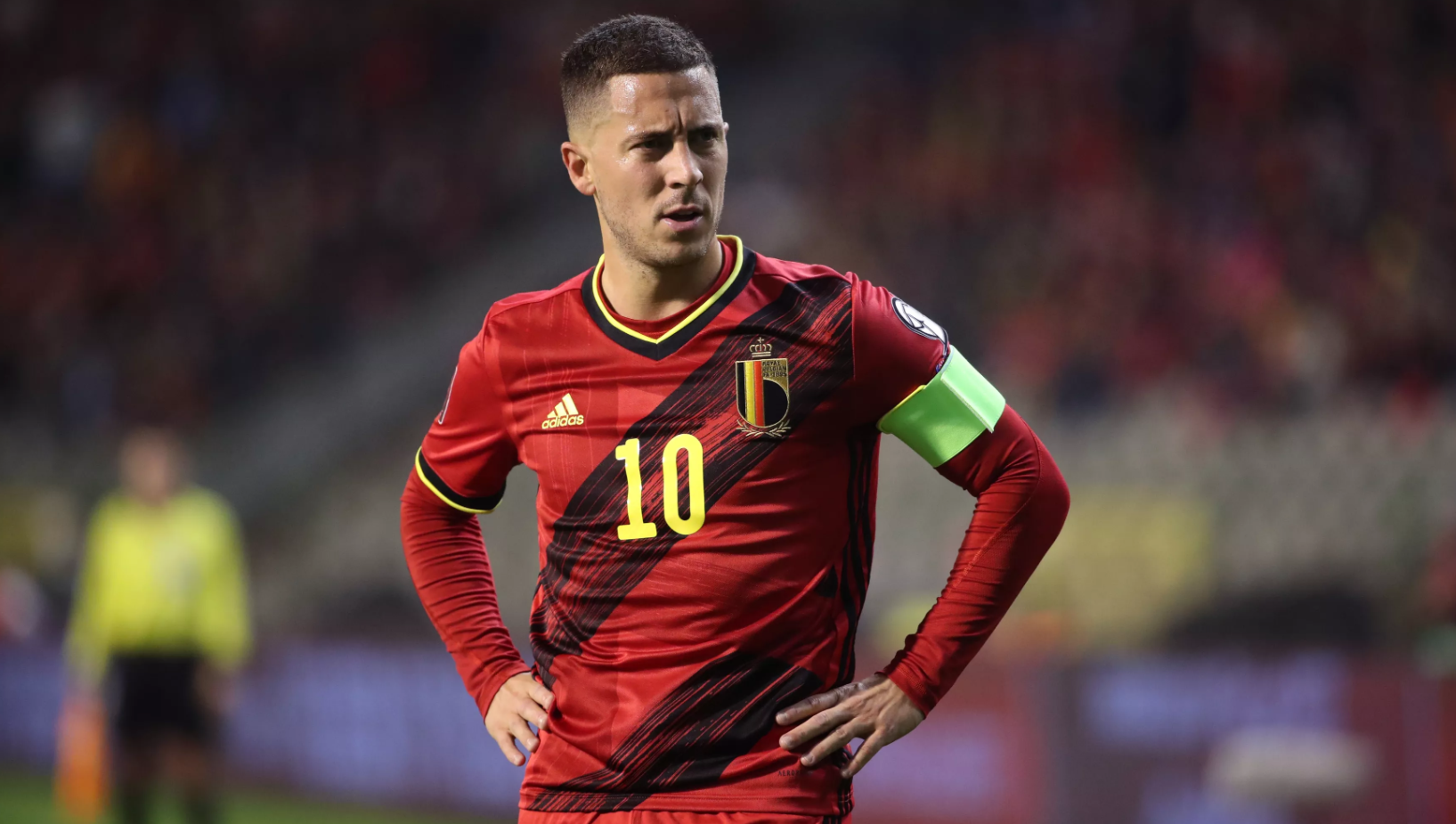 Eden Hazard u dresu belgijske reprezentacije u fudbalu crveni sa crnim dijagonalnim prugama dugi rukav drži ruke na kukovima broj 10 zelena kapitenska traka na terenu iza u bluru tribine namračen tamna kosa