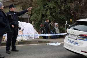 klasična sačekuša u Zagrebu policajac na ulici u uniformi pokazuje lijevom rukom na nešto u daljini drugom policajcu kapa na glavi ispred policijsko auto bijelo lijevo bijele vreće pored stoji troje djece u jaknama lica su im zamagljena bor oblačan dan