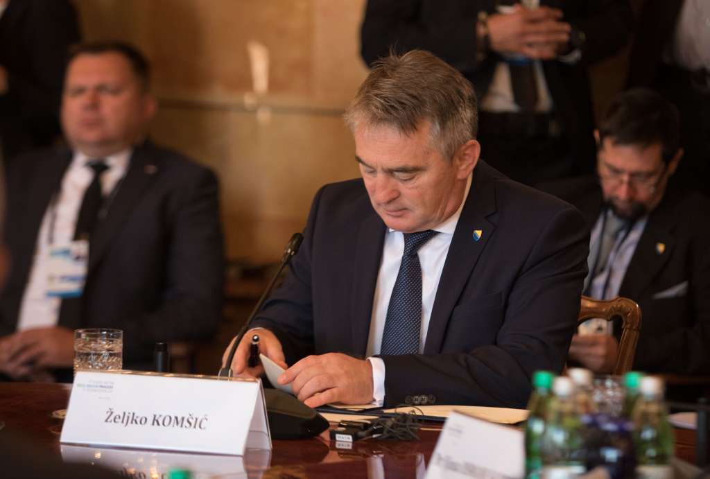 je lijepa vijest Željko Komšić sjedi u prostoriji ispred papiri drži olovku na stolu piše njegovo ime na bijelom papiru iza njega dva čovjeka u tamnom odijelu bijela košulja tamna šarena kravata voda na stolu